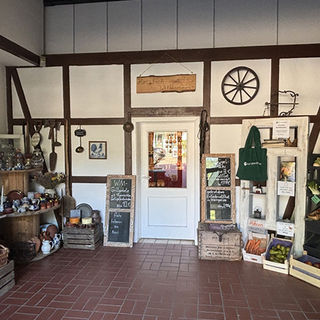Der Eingangsbereich des Hofladens auf Hof Bimöhlen ist im rustikalen Landhausstil gestaltet. Saisonale Blumen, historische Hofutensilien und dekorative Tafeln schaffen eine einladende Atmosphäre. Durch die Tür ist der Verkaufsraum mit regionalen Produkten sichtbar.