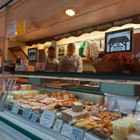 Das Bild zeigt den Marktstand von Hof Bimöhlen auf dem Wochenmarkt Eidelstedt. Hinter einer großzügigen Verkaufstheke mit Fleisch-, Wurst- und Feinkostprodukten stehen zwei Mitarbeiter und beraten die Kunden. Die Theke ist reichhaltig mit hausgemachten Spezialitäten und regionalen Erzeugnissen bestückt.