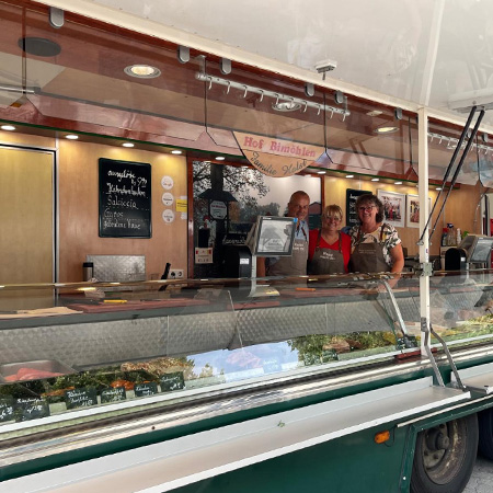 Das Bild zeigt den mobilen Verkaufsstand von Hof Bimöhlen auf dem Wochenmarkt Groß Flottbek. In der großzügigen Frischetheke werden verschiedene Fleisch-, Wurst- und Feinkostprodukte präsentiert. Eine Mitarbeiterin steht im Verkaufswagen bereit und berät Kunden zu den angebotenen regionalen Spezialitäten.