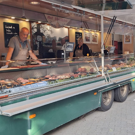 Das Foto zeigt die Verkaufstheke von Hof Bimöhlen auf dem Wochenmarkt Norderstedt. In der gekühlten Auslage werden verschiedene Fleisch-, Geflügel- und Feinkostprodukte präsentiert. Hinter der Theke stehen Mitarbeitende bereit, um Kunden zu beraten und regionale Spezialitäten anzubieten.